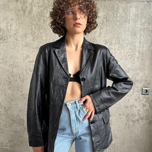 Puede incluir: Un blazer de cuero negro con solapa de muesca cl&aacute;sica y cierre de bot&oacute;n. El blazer se usa sobre un top negro y se combina con jeans azul claro. La chaqueta tiene un corte entallado y parece ser vintage.
