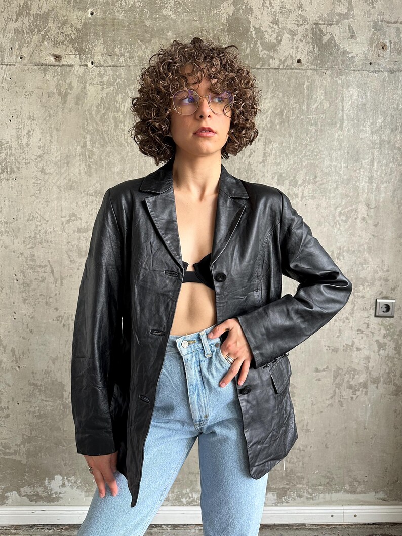 Puede incluir: Un blazer de cuero negro con solapa de muesca cl&aacute;sica y cierre de bot&oacute;n. El blazer se usa sobre un top negro y se combina con jeans azul claro. La chaqueta tiene un corte entallado y parece ser vintage.