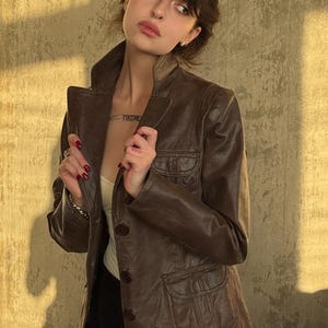 Peut inclure: Veste en cuir marron avec col classique et fermeture boutonnée. La veste comporte deux poches avant et une coupe ajustée. Le modèle porte un haut blanc et un pantalon noir. La veste est de couleur marron moyen.