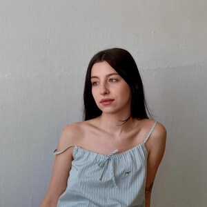 May include: A light blue and white striped camisole top with matching shorts. The top has thin straps and a small bow detail at the neckline. A small green embroidered design is visible on the top. The outfit is made of a lightweight fabric.