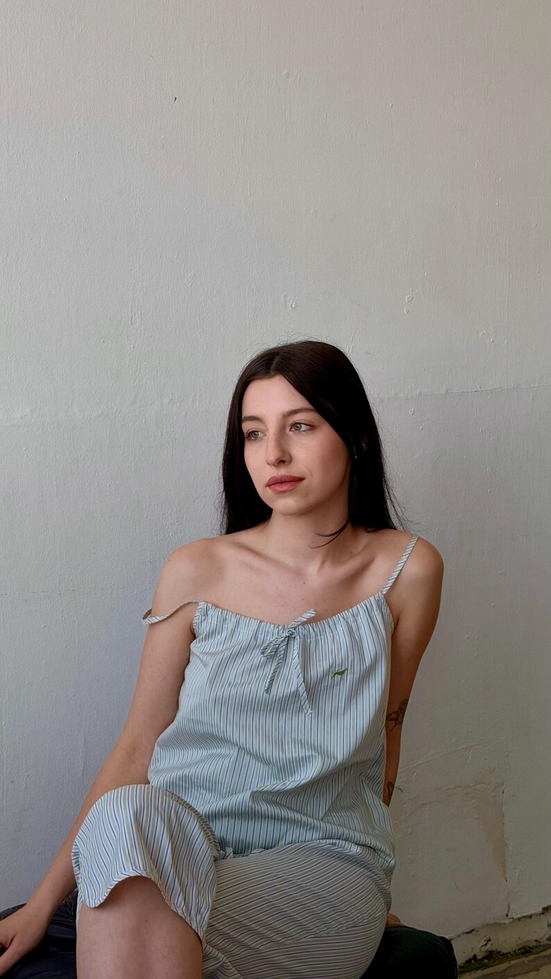May include: A light blue and white striped camisole top with matching shorts. The top has thin straps and a small bow detail at the neckline. A small green embroidered design is visible on the top. The outfit is made of a lightweight fabric.