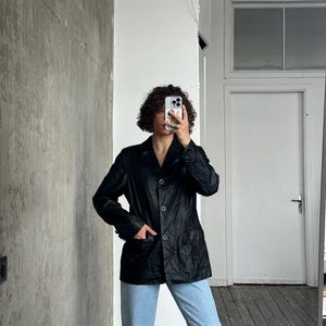 Puede incluir: Un blazer de cuero negro con un dise&ntilde;o cl&aacute;sico, con solapas de muesca, cierre de botones y bolsillos delanteros. El blazer se lleva con vaqueros azul claro. La imagen est&aacute; tomada en interiores, con una pared de tonos neutros en el fondo.