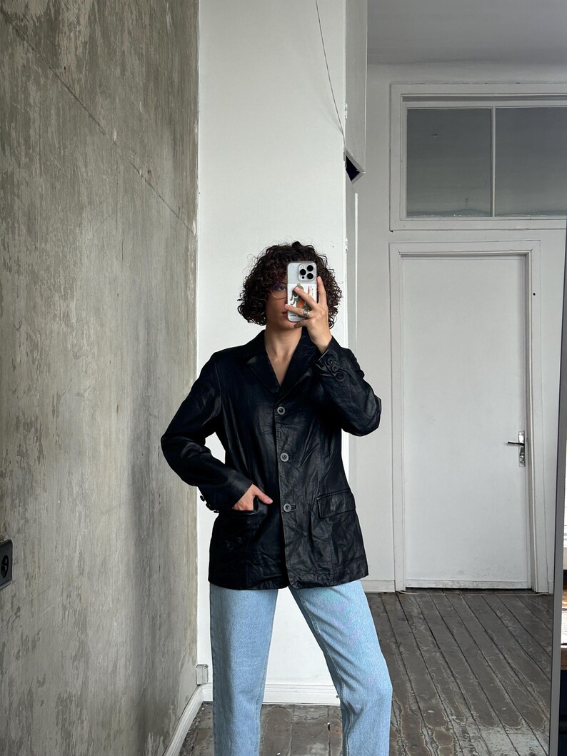 Puede incluir: Un blazer de cuero negro con un dise&ntilde;o cl&aacute;sico, con solapas de muesca, cierre de botones y bolsillos delanteros. El blazer se lleva con vaqueros azul claro. La imagen est&aacute; tomada en interiores, con una pared de tonos neutros en el fondo.