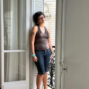 Op de afbeelding: Een bruine haltertop met een V-hals en geplooid stof bij de buste, gecombineerd met donkerblauwe denim knielange shorts met een knoop- en ritssluiting, en donkerblauwe schoenen met hak en enkelbandjes, gedragen met gelaagde armbanden.