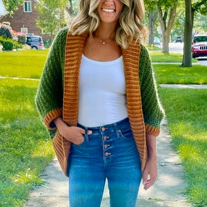 Dark green textured crochet shrug with gold folded over ribbed collar and cuffs, modelled on blond woman wearing white tank top and blue denim jeans, walking along a residential street.