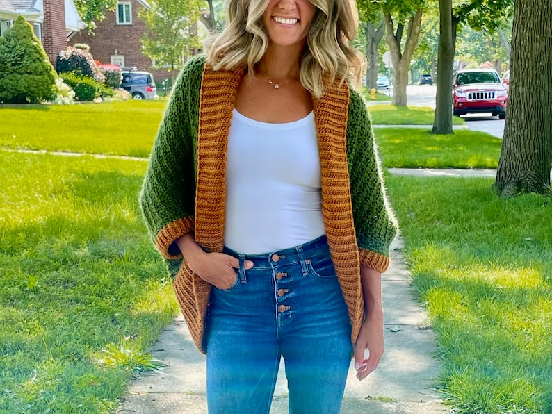 Dark green textured crochet shrug with gold folded over ribbed collar and cuffs, modelled on blond woman wearing white tank top and blue denim jeans, walking along a residential street.