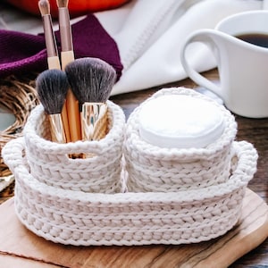 May include: A cream-colored, crocheted makeup organizer with two compartments. One holds makeup brushes, the other holds cotton pads. A white coffee mug, a black container with cinnamon sticks, and other decorative items are in the background.