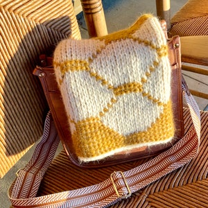 Folded cream and gold honeycomb crochet cowl laying on brown leather bag, sitting on wicker chair in the sunshine