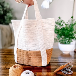 May include: A white and brown crocheted tote bag with a wide, flat base and two long handles. The bag is sitting on a wooden table with yarn and crochet tools.