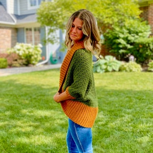 Dark green textured crochet shrug with gold folded over ribbed collar and cuffs, modelled on blond woman wearing white tank top and blue denim jeans, viewed in profile.