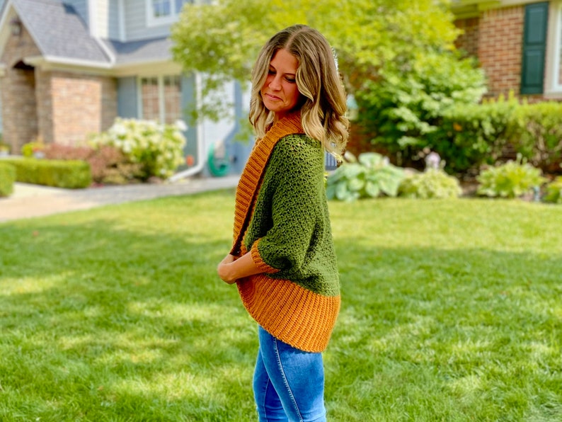 Dark green textured crochet shrug with gold folded over ribbed collar and cuffs, modelled on blond woman wearing white tank top and blue denim jeans, viewed in profile.