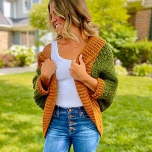 Dark green textured crochet shrug with gold folded over ribbed collar and cuffs, modelled on blond woman wearing white tank top and blue denim jeans, with one side off shoulder.