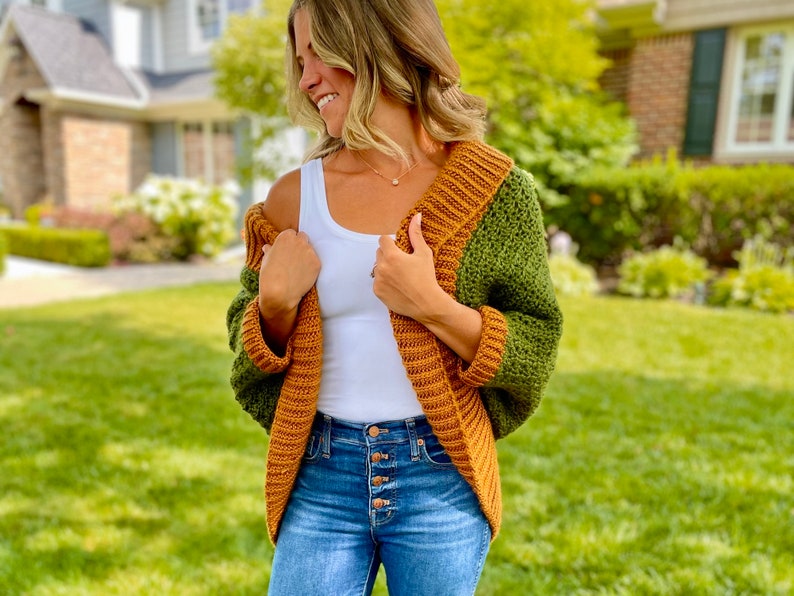 Dark green textured crochet shrug with gold folded over ribbed collar and cuffs, modelled on blond woman wearing white tank top and blue denim jeans, with one side off shoulder.