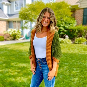 Dark green textured crochet shrug with gold folded over ribbed collar and cuffs, modelled on blond woman wearing white tank top and blue denim jeans.