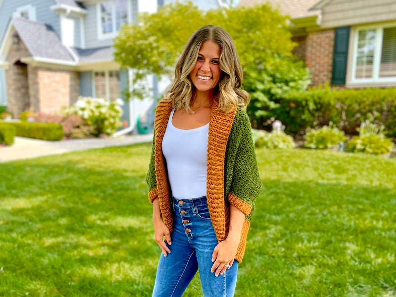 Dark green textured crochet shrug with gold folded over ribbed collar and cuffs, modelled on blond woman wearing white tank top and blue denim jeans.