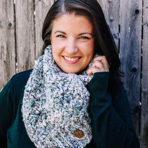 May include: A woman wearing a chunky knit infinity scarf in shades of gray, blue, and white. The scarf has two large wooden buttons and is paired with a dark green sweater. The background is a weathered wooden fence.