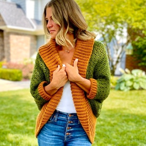 Dark green textured crochet shrug with gold folded over ribbed collar and cuffs, modelled on blond woman wearing white tank top and blue denim jeans.