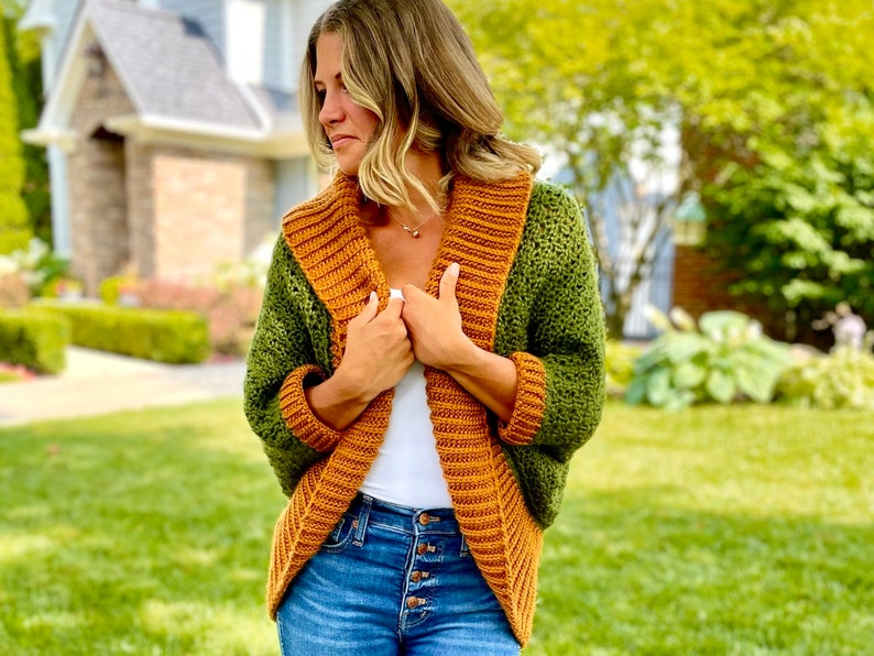 Dark green textured crochet shrug with gold folded over ribbed collar and cuffs, modelled on blond woman wearing white tank top and blue denim jeans.