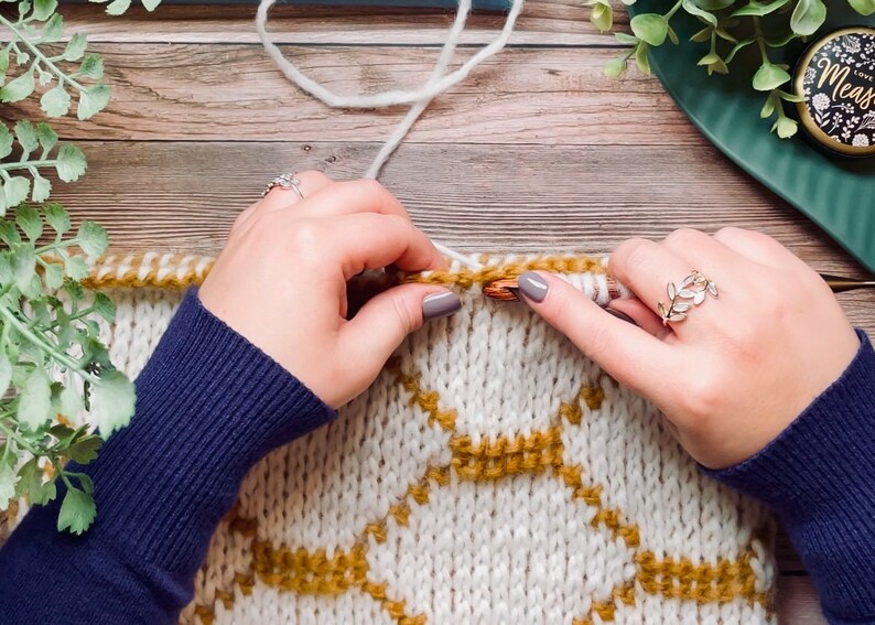 Close up tutorial of hands holding Tunisian crochet hooks and making a cream cowl with gold honeycomb motif, on a table.
