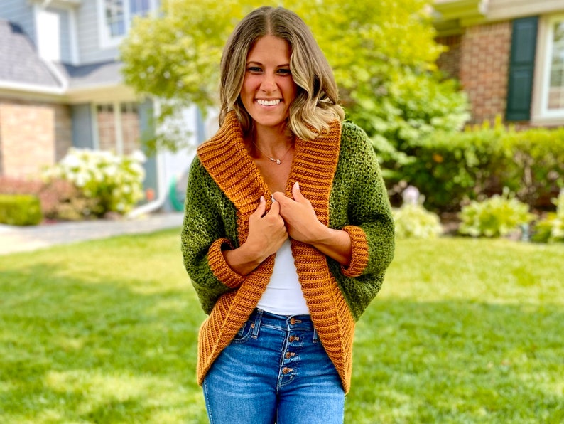 Dark green textured crochet shrug with gold folded over ribbed collar and cuffs, modelled on blond woman wearing white tank top and blue denim jeans.