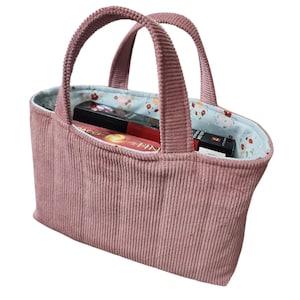 May include: A dusty rose corduroy tote bag with two matching handles. The interior is lined with a light blue fabric featuring a small floral pattern in red, orange, and green. A rectangular book with black and red spine text is inside the bag.