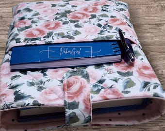 May include: A quilted fabric book cover featuring a floral pattern of pink roses and green leaves on a white background. The cover includes a front pocket holding a blue notebook with the text DakerGirl DESIGNS and a black pen. A tab closure secures the cover.