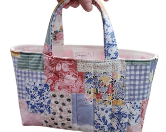 May include: A quilted fabric tote bag featuring a patchwork design of various floral and gingham patterns in shades of blue, pink, yellow, and white. The bag has two fabric handles and a light pink interior lining with a small white brand label.