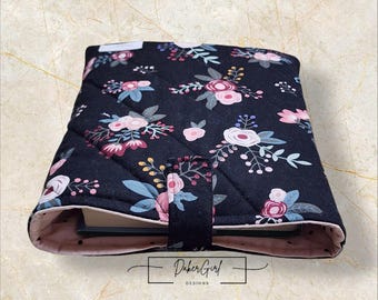 May include: A black quilted fabric book sleeve featuring a floral pattern with pink, white, and blue flowers and green leaves. The sleeve has a light pink interior lining and a central fabric tab closure. The text DakerGirl DESIGNS is visible.