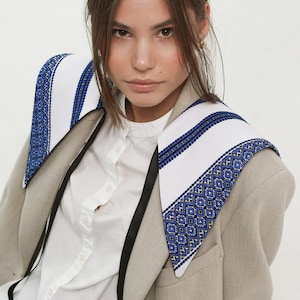 May include: A woman wearing a beige blazer with a white and blue embroidered collar. The collar has a black trim. She is also wearing a white button-down shirt and blue jeans.