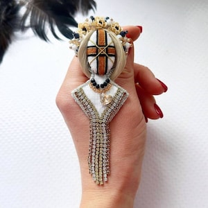 Op de afbeelding: Een witte stoffen broche met een gouden en zwarte kroon, een bruin en zwart kruisontwerp en een gouden en witte franje. De broche wordt in een hand met rode nagellak gehouden.