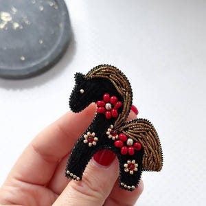 Könnte beinhalten: Schwarze Pferdebrosche mit roten Blumenakzenten und goldfarbenen Details. Mähne und Schweif des Pferdes sind mit goldfarbenen Perlen verziert. Ein einzigartiges Accessoire.