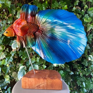 Könnte beinhalten: Eine farbenfrohe Holzskulptur eines Kampffisches mit einer blauen, grünen und orangefarbenen Schwanzflosse. Der Fisch ist auf einem Holzsockel montiert.