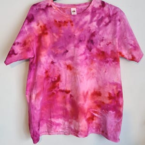 May include: A tie-dye t-shirt in shades of pink and red. The short-sleeved shirt has a crew neck and a vibrant, swirled pattern. The shirt is hanging on a black hanger against a white background.