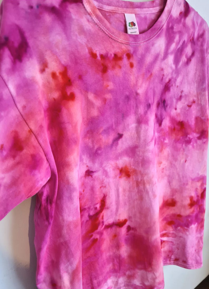 May include: A tie-dye t-shirt in shades of pink and red. The shirt has a crew neck and short sleeves. The tie-dye pattern is swirled and blended, creating a unique and colorful design.