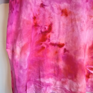 May include: A close-up of a pink tie-dye t-shirt. The shirt features a blend of vibrant pink and red hues, creating a unique, artistic pattern. The fabric appears soft and lightweight, with visible texture and a stitched hemline.