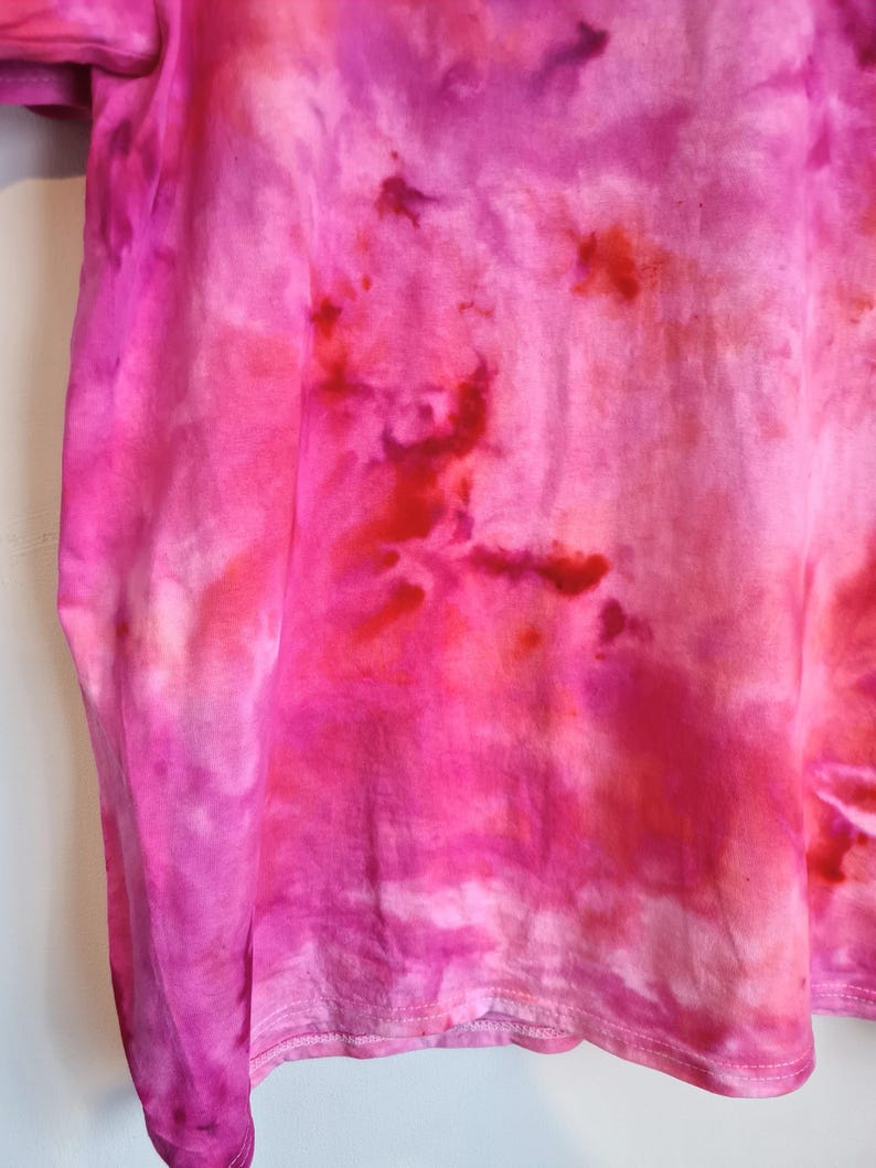 May include: A close-up of a pink tie-dye t-shirt. The shirt features a blend of vibrant pink and red hues, creating a unique, artistic pattern. The fabric appears soft and lightweight, with visible texture and a stitched hemline.