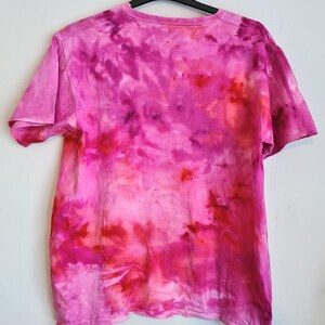May include: A short-sleeved, tie-dye t-shirt in shades of pink and red hangs on a black hanger against a white wall. The shirt's design features a blend of vibrant colors, creating a unique, artistic pattern. The fabric appears soft and comfortable.