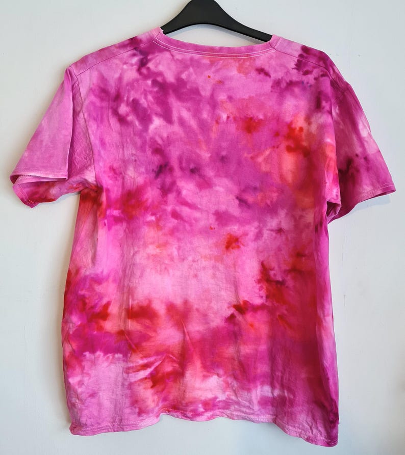 May include: A short-sleeved, tie-dye t-shirt in shades of pink and red hangs on a black hanger against a white wall. The shirt's design features a blend of vibrant colors, creating a unique, artistic pattern. The fabric appears soft and comfortable.
