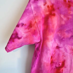 May include: Close-up of a tie-dye t-shirt in shades of pink and red. The short sleeve is visible, with a stitched seam and hem. The fabric has a soft, textured appearance, with a watercolor-like effect.
