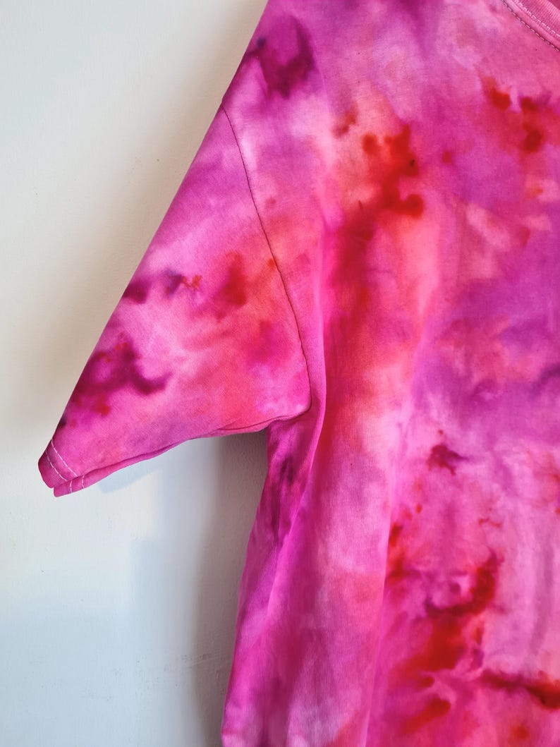 May include: Close-up of a tie-dye t-shirt in shades of pink and red. The short sleeve is visible, with a stitched seam and hem. The fabric has a soft, textured appearance, with a watercolor-like effect.