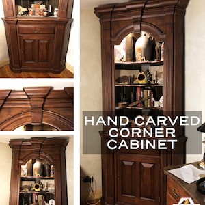 May include: A hand-carved corner cabinet with a dark wood finish. The cabinet has a glass door, shelves, and a lower cabinet door. The cabinet is decorated with intricate carvings. "HAND CARVED CORNER CABINET" is written on the front of the cabinet.