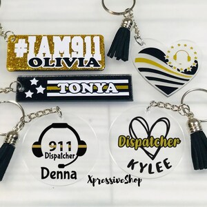 May include: A collection of acrylic keychains featuring personalized names and 911 dispatcher themes. Designs include rectangular, heart, and circular shapes with gold glitter, black, white, and yellow accents, each attached to a metal ring and tassel.