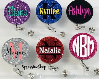May include: Six glitter badge reels with different names and monograms. The names are Eliana, Kynlee, Ashlyn, Megan, Natalie, and NBM. The reels are all round and have a clear plastic strap.