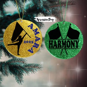 May include: Two glittery round ornaments. One is gold with a black flag silhouette, the name "AMARA" in blue, and the year "2025". The other is green with two black flags and the word "HARMONY". Both have hanging strings.