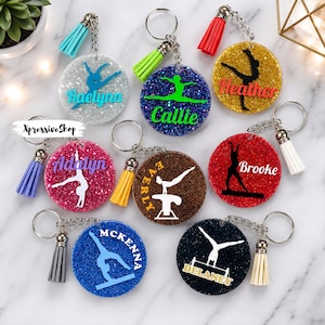 May include: Eight circular glitter keychains with metal rings and colorful tassels. Each keychain features a gymnast silhouette and a name in vinyl text. Names include Raelynn, Callie, Heather, Adalyn, Everly, Brooke, Mckenna, and Delaney. Text XpressiveShop is visible.