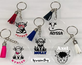 May include: Five circular clear acrylic keychains with metal rings and colorful tassels. Each features a unique black graphic of a highland cow or bull, with names printed in various colors including red, blue, pink, and white. Text reads XpressiveShop.