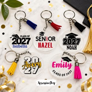 May include: Five circular clear acrylic keychains with metal rings and colorful tassels. Each features graduation-themed text and graphics, including names like Isabella, Hazel, Noah, Avery, and Emily, along with the years 2021 or 2027 and graduation caps.