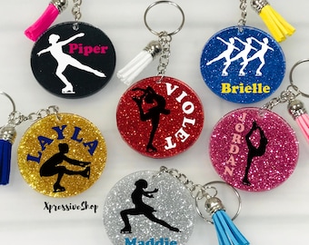 May include: Six circular glitter keychains featuring ice skater silhouettes and names. Colors include black, red, blue, gold, pink, and silver. Each keychain has a metal ring and a small tassel. Names include Piper, Violet, Brielle, Layla, Jordan, and Maddie.