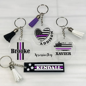 May include: Five different keychain designs with a purple thin line on a black and white American flag design. The keychains are made of clear acrylic and have a metal ring. The names Addison, Brooke, Xavier, and Kendall are printed on the keychains. The keychains have black, white, and purple tassels.