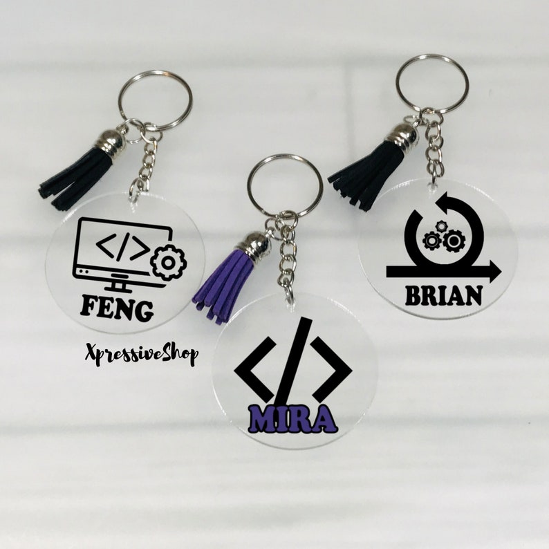 Peut inclure: Trois porte-cl&eacute;s en acrylique transparent avec des ic&ocirc;nes et des noms noirs et blancs. Le premier porte-cl&eacute;s repr&eacute;sente un &eacute;cran d'ordinateur avec du code &agrave; l'&eacute;cran et le nom "FENG". Le deuxi&egrave;me porte-cl&eacute;s repr&eacute;sente une fl&egrave;che circulaire avec des engrenages et le nom "BRIAN". Le troisi&egrave;me porte-cl&eacute;s repr&eacute;sente des symboles de code et le nom "MIRA". Chaque porte-cl&eacute;s est dot&eacute; d'un pompon violet.